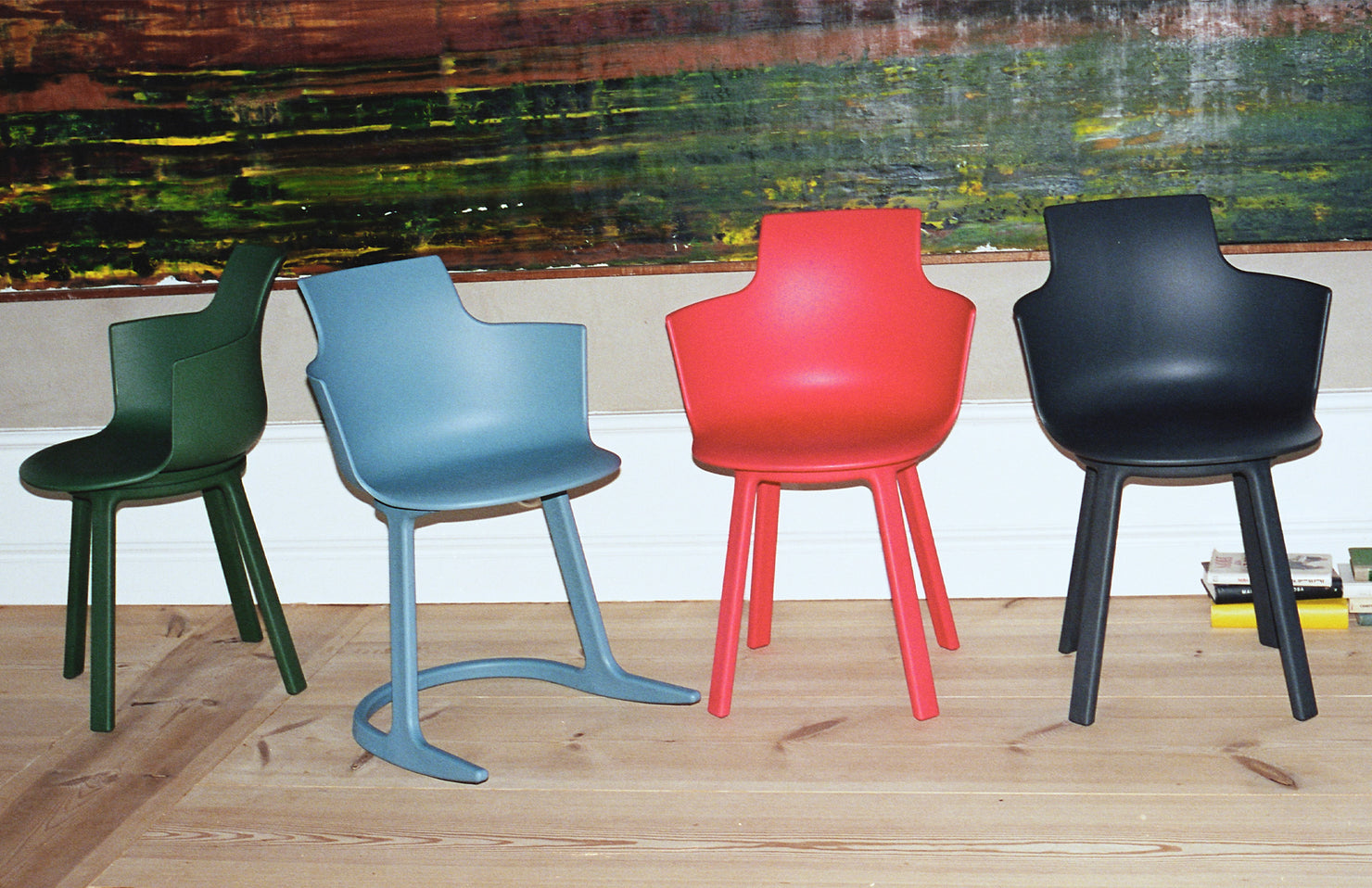 Four colorful chairs (green, blue, red, black) arranged in a row on a wooden floor with a painting in the background.